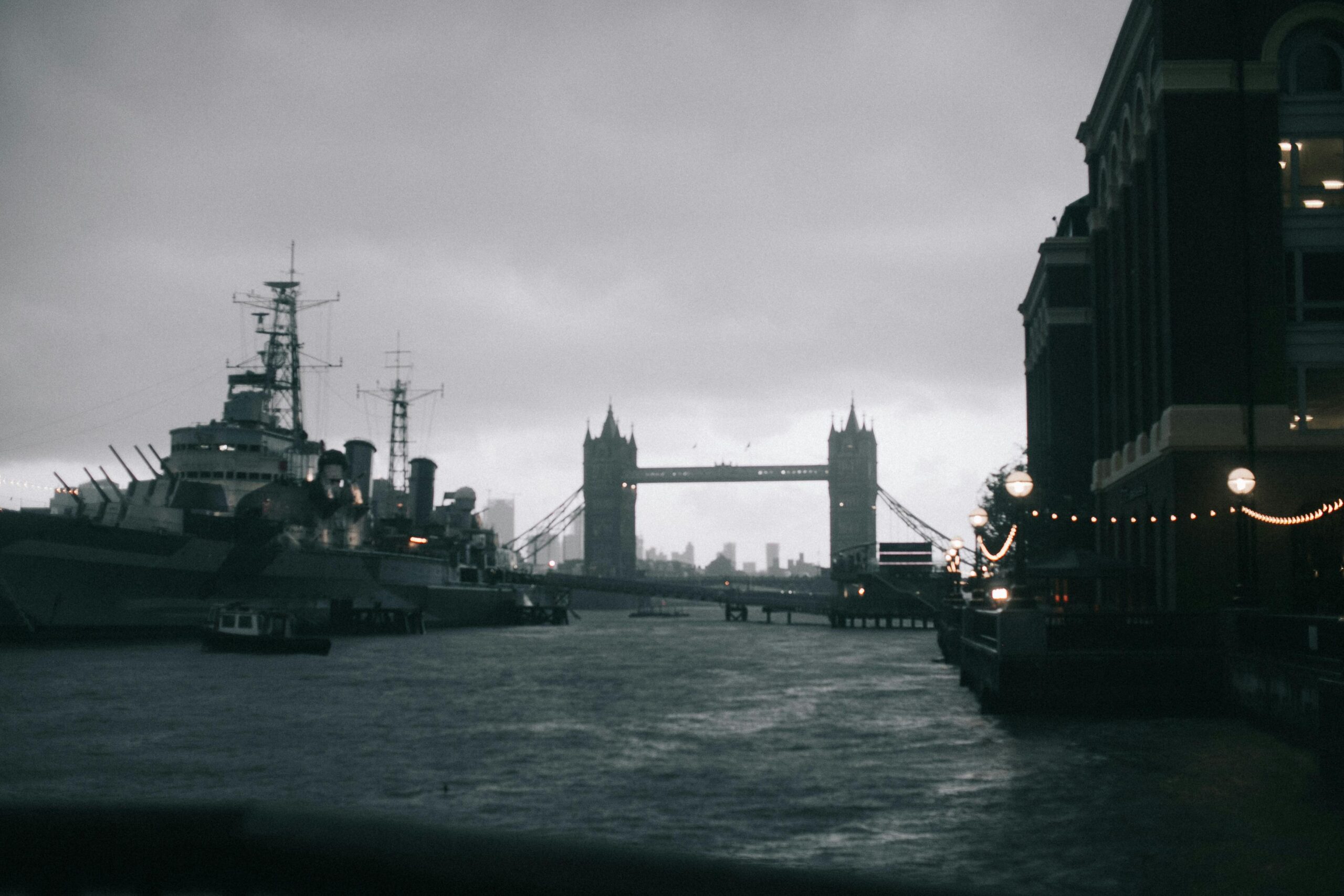 Dramatic black and white photo of Tower Bridge in London with warship and water reflections at dusk.