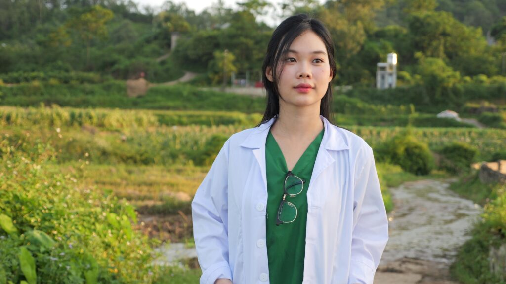 A young female healthcare professional stands outdoors with greenery in the background.