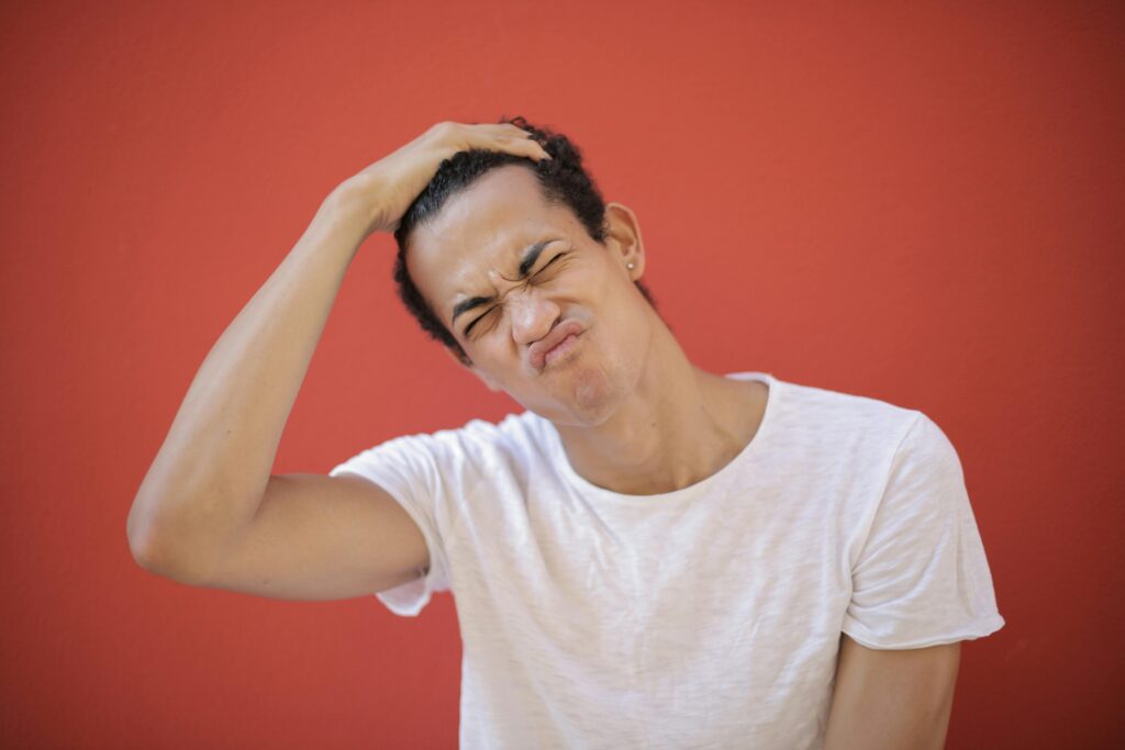 Young man making a funny face, scratching head against a red backdrop, showing playful expression.