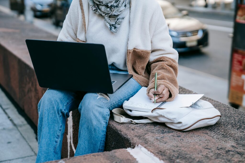 Unrecognizable woman multitasking outdoors with laptop and notebook on a city bench.