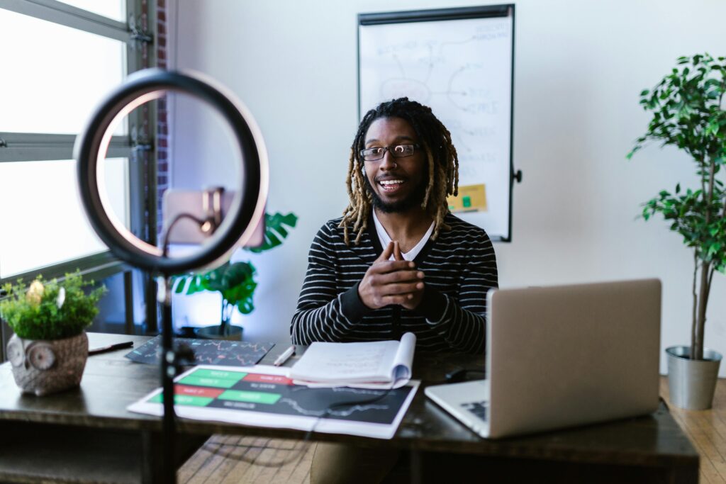 Young man with dreadlocks creating a video blog using smartphone and ring light in home office.