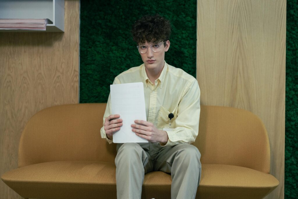 Young man sitting on a sofa in an office holding papers, wearing glasses and a yellow shirt.