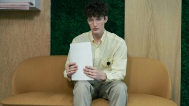 Young man in yellow shirt and glasses sits on sofa in an office, holding papers.